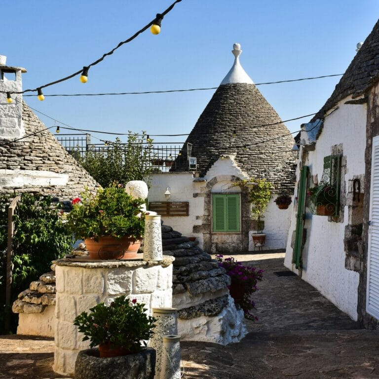 A stunning view of the Trulli of Alberobello, showcasing the iconic circular limestone houses with conical roofs, surrounded by cobblestone streets and vibrant greenery under a bright blue sky.